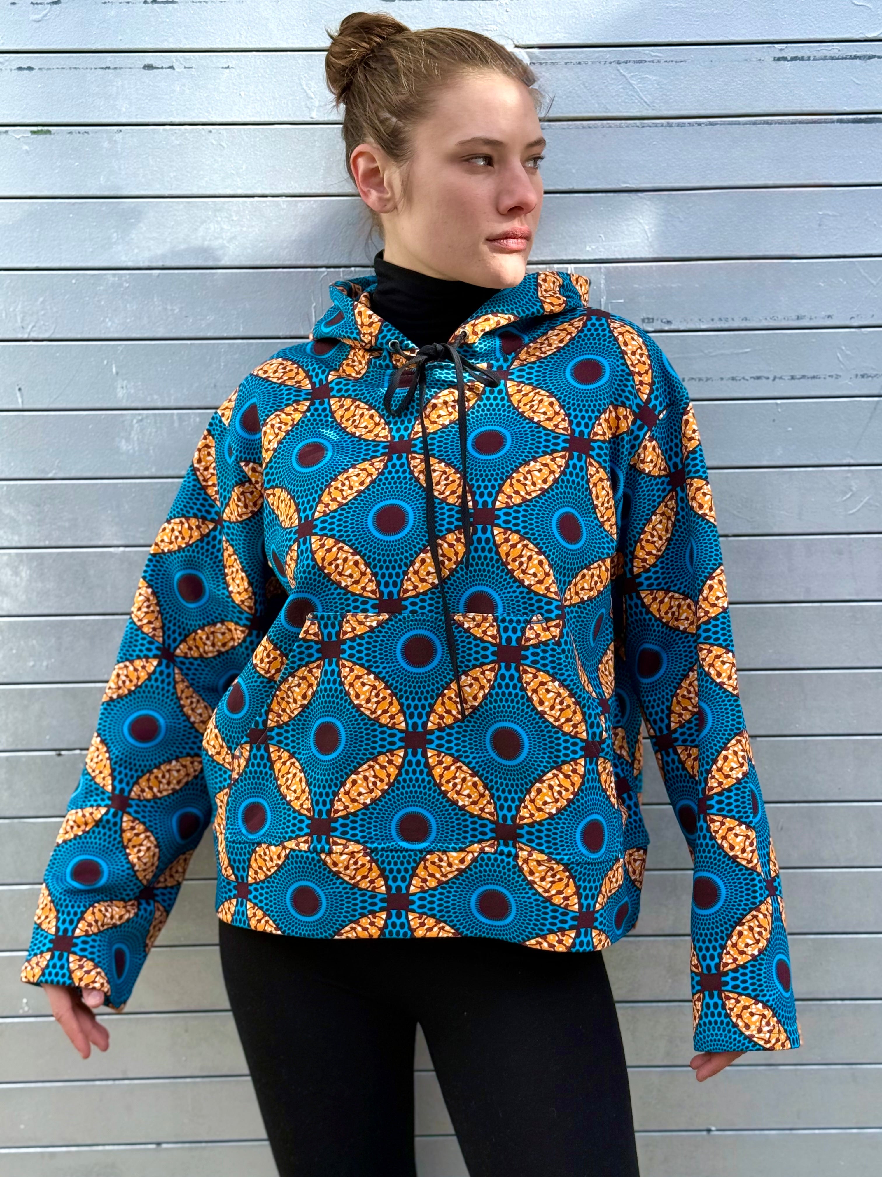 Model in the Ìmọ́lẹ̀ Blue Sweat Hoodie by Lola Faturoti Loves, looking away under soft daylight. The circular Yoruba motifs in blue and yellow evoke balance, strength, and inner light through wearable art.